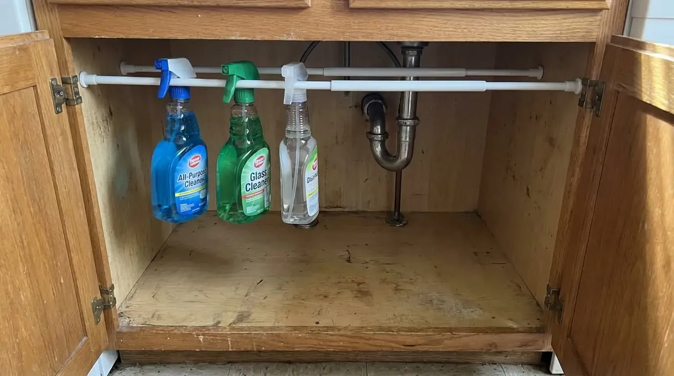 Three spray bottles of cleaning solution hanging by their triggers from a white tension rod inside a wooden under-sink cabinet.