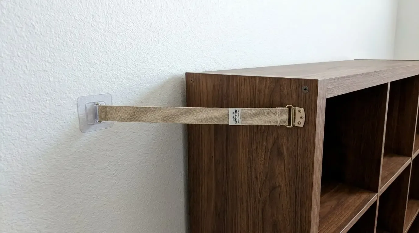 Beige heavy-duty anti-tip safety strap anchored from a white wall to a wood shelf.